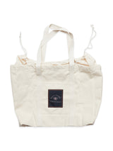 The BDG Canvas Bag - Off White by Blue de Gênes is an everyday beige tote featuring two sturdy handles and a drawstring closure. A black patch with white text and a turtle image adds whimsy to this practical accessory.