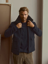 A man with light hair and a beard stands indoors against a neutral backdrop, wearing the Blue de Gênes BGdugal Bomber Jacket in Dark Navy over a dark sweater and light corduroy pants, holding his jacket collar with both hands.