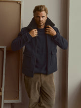 A man with blonde hair and a beard stands indoors, adjusting his navy Blue de Gênes jacket worn over a blue sweater. He wears Blue de Gênes BGpalco Cord Chino pants in Sepia Tint Brown, with framed canvases leaning against the wall behind him.