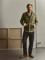 A man stands indoors wearing a green jacket, Blue de Gênes BGmarco Connor Jeans in Black Denim Used—crafted from 100% cotton denim—brown boots, and a scarf. He faces forward with his hands in his pockets while a blank canvas leans against the wall behind him.