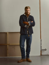A man standing indoors with arms crossed, wearing a navy jacket, plaid shirt, Blue de Gênes BGgiacomo BB Jeans in blue black denim, brown boots, and orange gloves, positioned in front of a plain wall and a wooden frame.