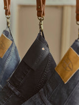 A close-up shows three pairs of jeans suspended by leather straps. The prominently displayed pair is the BGvinci TrueBlack Jeans in black denim from Blue de Gênes, featuring a label with the brand’s name and a small shield logo. The other jeans are blue regular fit styles with brown leather patches, all made from comfort stretch denim for added ease and flexibility.
