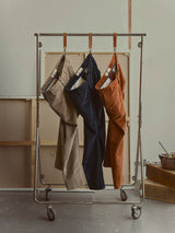 A pair of BGpalco Cord Chino trousers in Sepia Tint Brown by Blue de Gênes hangs neatly from a leather strap on a metal clothing rack in a minimalist studio, surrounded by canvases and wooden boxes, highlighting the brand’s signature tapered silhouette.
