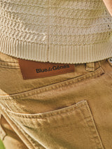 Close-up of a person wearing BGgiacomo Color Jeans - Tanin by Blue de Gênes in light brown corduroy, featuring a leather brand patch, paired with a cream knit sweater partially tucked into the waistband.