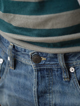 Close-up of a person in Blue de Gênes BGgiacomo Tim Jeans - Antique wash, featuring blue denim, relaxed fit, yellow stitching, classic waistband details, and logo button. The outfit is paired with a green and gray striped shirt.
