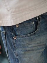 Close-up of a person in Blue de Gênes BGgiacomo Tim Jeans - Antique wash, made in Italy. The relaxed fit jeans show a red cross emblem on the pocket, with distinct stitching and textured fabric clearly visible.