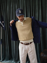 A man in a navy baseball cap, yellow striped polo, cream trousers with the Blue de Gênes BGcollege Belt - Green Stripe, and a navy jacket stands before dark green curtains, putting on the jacket. Flags can be seen in the background.