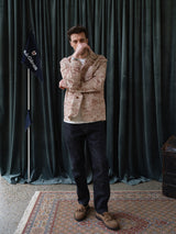 A man stands indoors on a patterned rug by dark green curtains, wearing the Blue de Gênes BGkendrick Print Jacket - Multi Pattern over a white shirt, with dark jeans and brown shoes, partially covering his face. A dark flag and wooden chest are nearby.