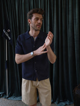 A man stands indoors in front of dark green curtains and a flag, wearing a navy short-sleeve button-up shirt, Blue de Gênes BGcopple Rodas Shorts in Pale Khaki, and a wristwatch, with his hands together as if adjusting his wrist.