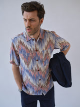 A man wears a short-sleeve button-up shirt with a colorful zigzag pattern and Blue de Gênes BGmenzino Vecchio Shorts in dark navy, holding a navy jacket over his left arm and looking slightly left against a plain light background.