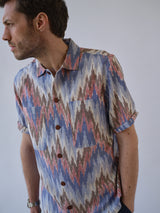 A man wears the Blue de Gênes BGponti Kala S/S Shirt in a bold multicolor pattern, featuring zigzag and tribal-inspired designs, against a plain light background, looking to his left.