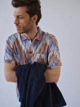 A man with short brown hair wears a Blue de Gênes BGponti Kala S/S Shirt - Multi Pattern, holding a dark jacket and looking to the side against a plain light background.