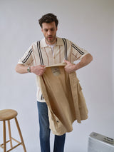A man showcases the BGhunter Vecchio Jacket in Pale Khaki by Blue de Gênes indoors, holding it open to reveal the interior. He pairs it with a striped short-sleeve shirt and blue jeans, next to a wooden stool and silver suitcase on a light background.