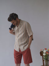 A man stands indoors looking down, wearing a beige short-sleeve striped shirt and Blue de Gênes BGfloyd Cord Shorts - Coral. He holds a jacket over his shoulder. Beside him, a wooden chair with a vase of colorful flowers sits against a plain background.