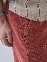 A close-up of a person wearing red corduroy pants, a Blue de Gênes BGcody Fling Shirt in Pale Khaki with dark vertical stripes and relaxed fit, plus a silver wristwatch and shield-shaped patch near the pocket.