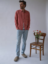 A man wears the Blue de Gênes BGenric Hope L/S Shirt - Coral, light blue jeans, and beige shoes, standing next to a wooden chair with a glass vase of colorful flowers against a plain light background.