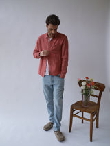 A man in a red button-up shirt, Blue de Gênes BGvinci Trueblue Jeans - Mid Blue Denim, and beige shoes stands looking down beside a wooden chair with a glass vase of colorful flowers, set against a plain light background.