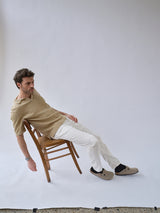 A man in the Blue de Gênes BGjohn Slubby Polo in Pale Khaki and white pants leans back on a wooden chair, one arm down. He wears beige slippers and black socks against a plain white backdrop.