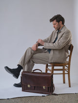 A man wearing the Blue de Gênes BGjazzy Ola Jacket in Dark Sand and black shoes sits on a wooden chair, checking his watch. A large brown leather briefcase is at his feet against a plain white backdrop.