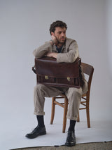 A man wears the Blue de Gênes BGjazzy Ola Jacket in Dark Sand, sitting on a wooden chair with his elbow on a brown leather briefcase. He pairs it with black shoes and dark socks, gazing thoughtfully to the side against a plain background.