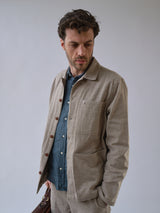 A man with wavy hair wears the Blue de Gênes BGenric Hope L/S Shirt in Green Harvest under a light beige jacket, holding a brown bag and looking down to his left against a plain light background.