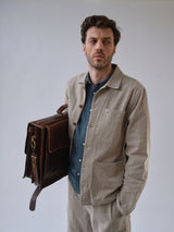 A man with short brown hair wears the Blue de Gênes BGjazzy Ola Jacket in Dark Sand over a blue shirt, paired with light beige pants, and stands against a plain background holding a large brown leather briefcase on his shoulder.