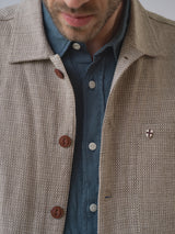 A man wears a textured beige jacket with brown buttons and a shield-shaped pin over the Blue de Gênes BGenric Hope L/S Shirt - Green Harvest. The image partially shows his face, focusing on the outfit.