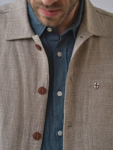 A man wears the BGjazzy Ola Jacket - Dark Sand by Blue de Gênes, textured beige cotton linen with brown buttons and a small shield-shaped pin, over a blue collared shirt. Only his lower face and chest are visible.