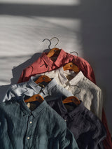 Five Blue de Gênes BGenric Hope L/S shirts in chalk, red, blue, gray, and dark green hang on wooden hangers against a sunlit white wall—striped shadows highlight their breathable linen for the perfect summer shirt display.