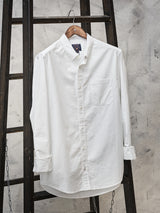A Sandro Oxford Shirt - White by Blue de Gênes, made from organic cotton with a front pocket, hangs on a wooden hanger. Its set against a dark wood ladder background, draped with chains for a rustic, industrial vibe.