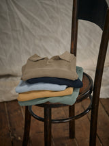 A stack of Filo Knit Polo shirts by Blue de Gênes in beige, navy, light blue, mustard, and sage rests on a dark-framed wooden chair. Made from 100% cotton, they sit on a wooden floor against a light fabric background.