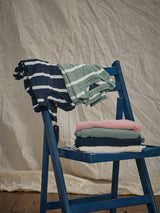A blue folding chair holds folded clothes, with a navy and white striped shirt on top. Below, theres a mint green shirt, Blue de Gênes Monte Knit in Porcelain Pink, and a dark green shirt made from soft cotton/linen blend. A textured white fabric drapes in the backdrop for a serene scene.