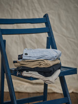 A stack of Menzino Shorts in light blue, beige, black, and khaki rests on a bright blue wooden chair against a beige cloth backdrop. Crafted by Blue de Gênes, the collection includes garment-dyed pieces and herringbone fabric options for added texture.