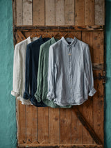 Four Enrico Beach Shirts by Blue de Gênes, in white, navy, green, and light blue hang on wooden hangers against a rustic door. Corozo nut buttons accent each piece, set against a teal wall backdrop, adding contrast and texture to this perfect summer shirt scene.