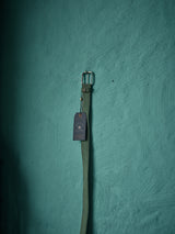 The Suede Belt - Green Bay by Blue de Gênes, featuring a silver buckle, hangs on a textured teal wall. A dark tag near the buckle adds sophistication to the display. Handmade in Italy, this accessory beautifully blends artistry with tradition.