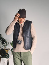 A man wearing a brown knit beanie, beige sweater, and the Blue de Gênes Saga Shearling Gilet - Scarab stands indoors with one hand on his head, a camera around his neck, and a potted plant beside him.