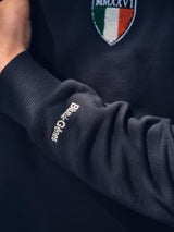 A close-up of a person’s arm wearing the BGclubhouse Rugby Sweatshirt - Navy by Blue de Gênes, with embroidered branding on the sleeve and an Italian flag patch marked MMXXVI on the chest.