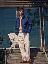 A man stands on wooden steps outdoors, wearing the Blue de Gênes BGcanopy Jacket in Navy Peony, paired with light beige pants, a striped shirt, and brown shoes. Behind him is a building with large glass windows and a white outdoor chair.