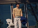A man stands outside large glass windows wearing the Blue de Gênes BGbob Knit Polo in Sunflower and white pants, holding a blue jacket. A white chair behind him completes his relaxed, nautical look.