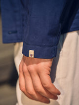 A close-up of a hand resting by the side, wearing the Blue de Gênes BGcanopy Jacket in Navy Peony with a beige tag and red-and-blue shield emblem at the cuff, paired with light-colored pants.