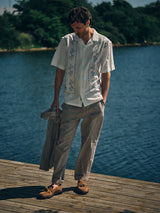 A man stands on a dock by a lake, holding the Blue de Gênes BGjazzy Ola Jacket in Dark Sand. He wears a short-sleeved, white embroidered shirt, light brown trousers, and brown boat shoes with trees in the background.