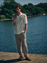 A man stands on a wooden dock by the water in a white embroidered cotton-linen shirt, beige pants, and brown shoes. He has the Blue de Gênes BGjazzy Ola Jacket in Dark Sand draped over one shoulder, surrounded by lush green trees.