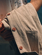 A person holds the BGjazzy Ola Jacket in Dark Sand by Blue de Gênes, featuring brown buttons and textured fabric, draped over their arm. In the background, a shirt with an embroidered floral design is partially visible.