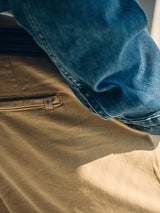 Close-up of a person wearing Blue de Gênes BGpalco Rodas Pants in Pale Khaki, featuring a relaxed fit, visible back pocket with fabric tag, paired with a dark belt and blue denim jacket. Sunlight highlights these smart-casual chinos.