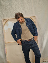 A man in a 100% cotton khaki Filo Knit Polo by Blue de Gênes, wearing a blue denim jacket and jeans, gazes downward. He stands before a large blank canvas set against a draped cloth background.