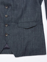 A close-up of a Blue de Gênes Casella Wasabi Blazer in denim blue, featuring a front pocket and five Buffalo horn buttons on the left side. The fabric showcases a subtle textured pattern with a small opening at the bottom of the jacket.
