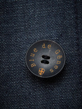 A close-up of a dark Buffalo horn button on blue denim fabric shows Blue de Gênes and a crest logo, echoing the craftsmanship of the Casella Wasabi Blazer by Blue de Gênes.