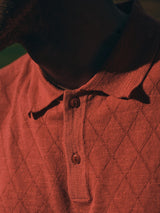 Close-up of a person wearing the Blue de Gênes BGwally Diamond Polo in Coral, featuring a diamond-knit pattern and two fastened buttons. Warm lighting highlights the lower face and neck, partially visible in shadow.