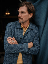 A man with brown hair and a mustache stands with arms crossed, wearing a blue patterned jacket over a Blue de Gênes Filo Knit Polo in Yellow Grain. Hes looking to the side, leaning against a dark teal fabric backdrop, with his ring visible on his hand.