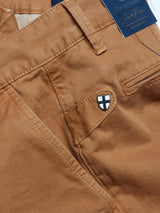 Close-up of Franco Genes Chino - Biscotto by Blue de Gênes: brown with side pocket detail, featuring a small black and white shield emblem on the pocket flap. The fabric tag showcases elegant script and decorative elements, highlighting the garment-dyed craftsmanship.